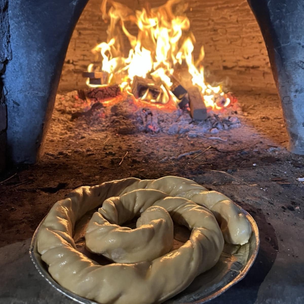 Παραδοσιακή πίτα δίπλα σε ξυλόφουρνο με αναμμένη φωτιά, που αποδίδει αυθεντική αίσθηση ψησίματος.