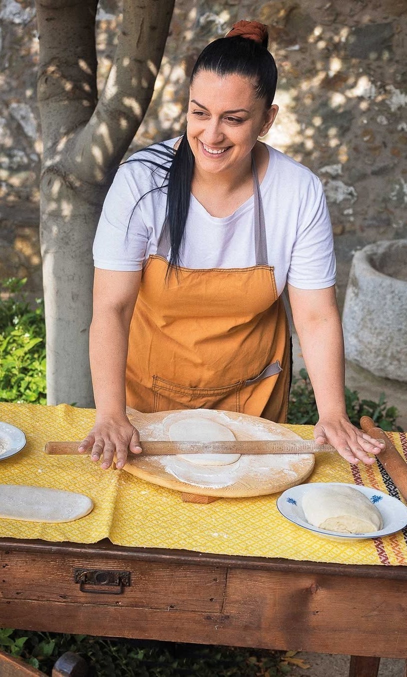 Χαμογελαστή τεχνίτρια δίπλα σε παραδοσιακό τραπέζι εργασίας με ζύμη και σκεύη παρασκευής.